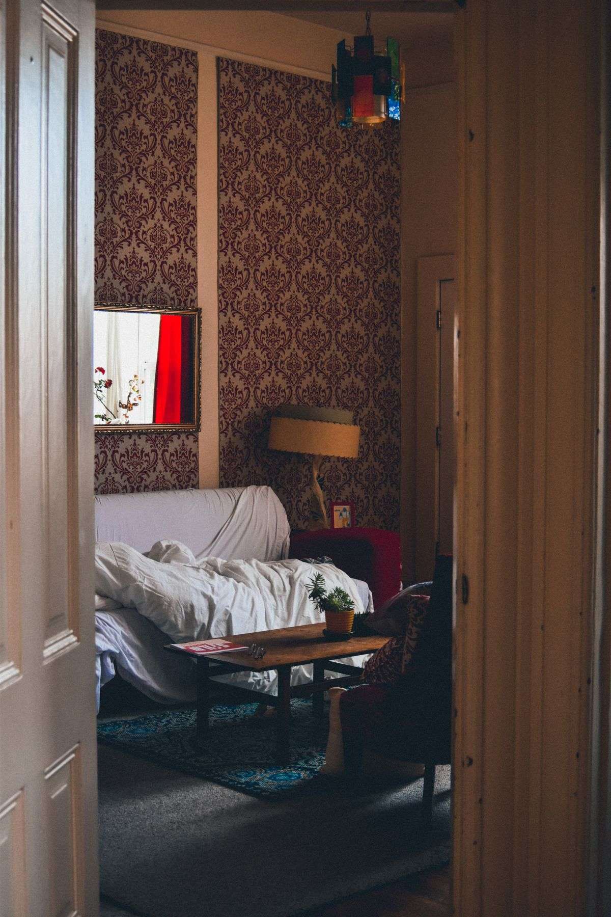 A view through a half-open door into a lived-in sitting room with patterned wallpaper, a lamp and a couch